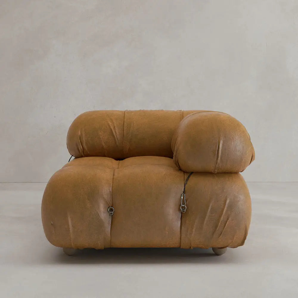 The Belmont Sofa Left Armrest features plush tan leather, rounded cushiony shapes, and visible connecting straps, evoking iconic classic style against a light beige background.