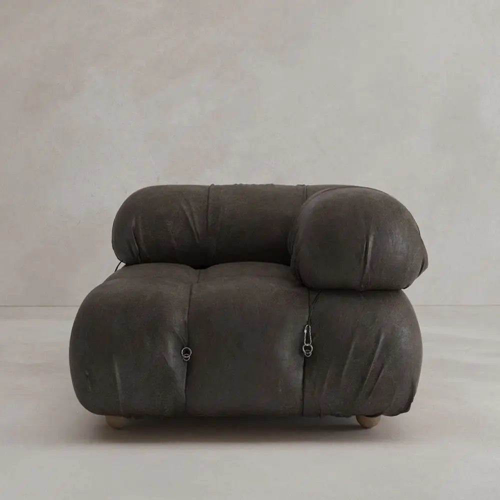 The Belmont Sofa Left Armrest features a modern, oversized design in dark gray with rounded, tufted cushions and visible metal fasteners, highlighting its modular style against a plain, light wall and floor.