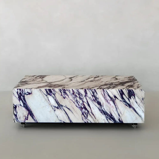 The Calacatta Viola Coffee Table features striking purple and black veining on a rectangular marble top, set on small wheels. Displayed against a light gray background, it adds sophisticated style to any room.