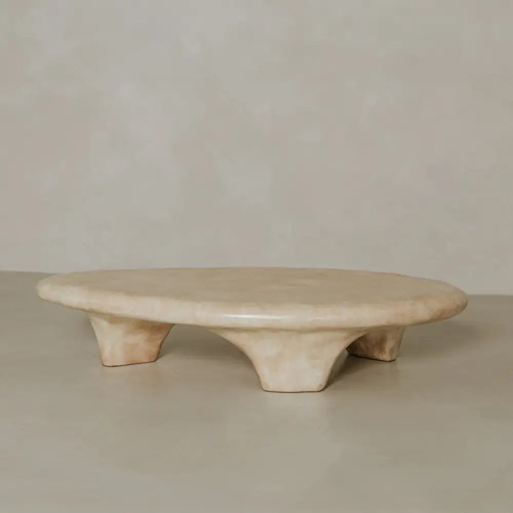 The Cleo Coffee Table features a low, oval beige stone platform with three wide curved legs showcasing brutalist design, set on a smooth neutral surface against a light textured background.