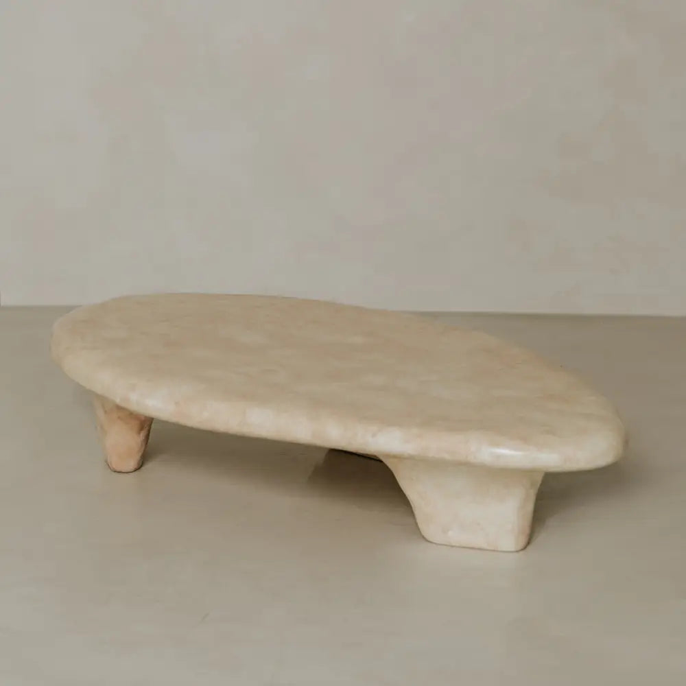 The Cleo Coffee Table features an oval, beige stone design with two short, wide legs, set against a matching beige wall and floor. Its smooth, polished surface and minimalist form subtly reference brutalist style.