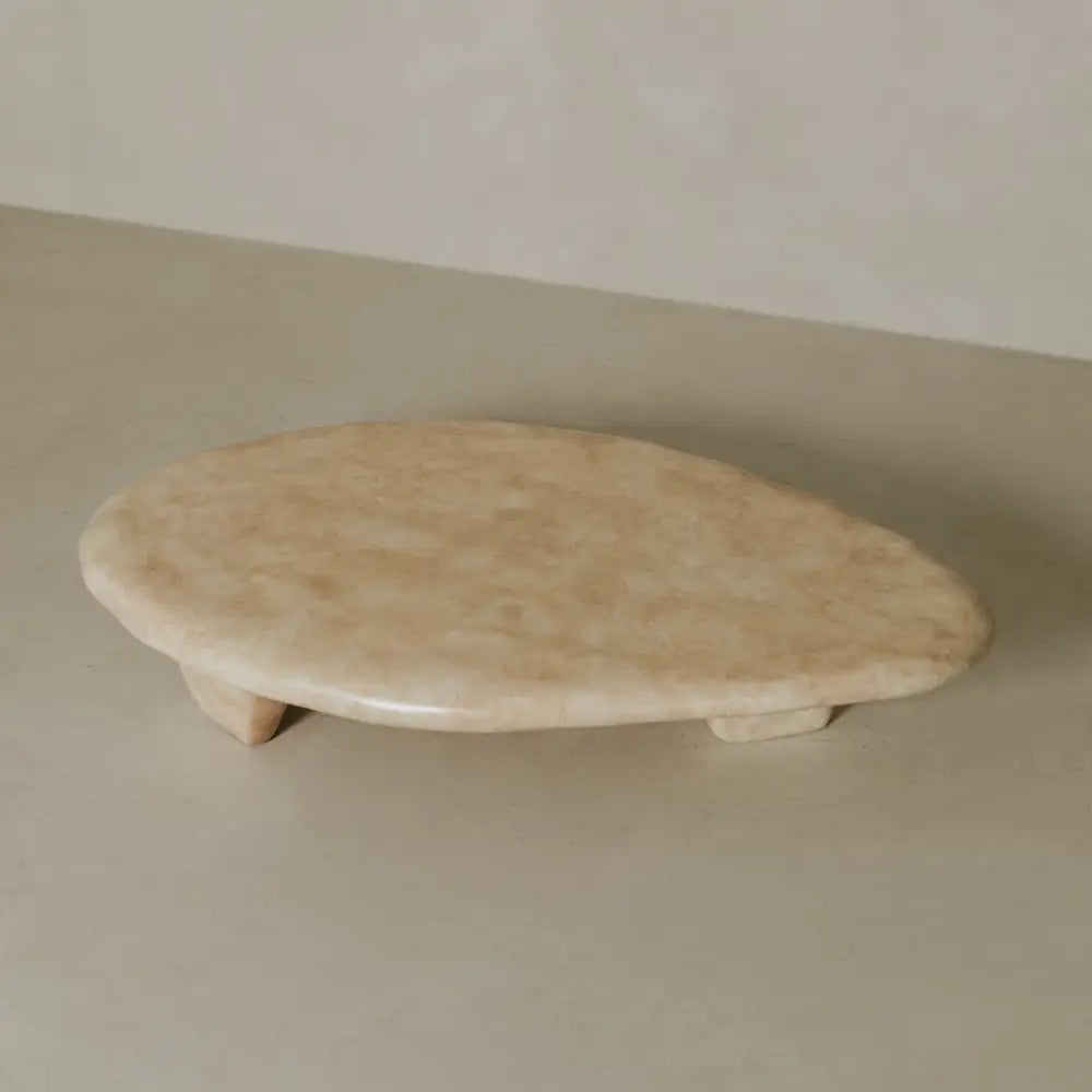 The Cleo Coffee Table features a smooth, oval beige stone top with short legs, showcased on a neutral surface and set against a light, textured background.