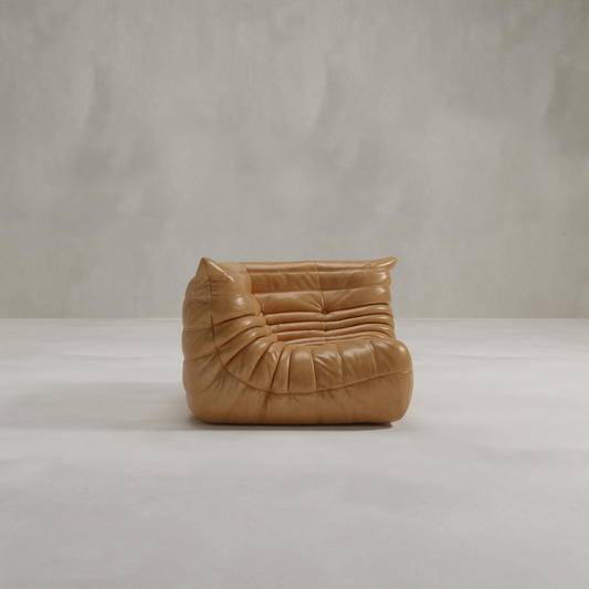 The Rolo Lounge Corner Sofa, a modern tan leather piece with deep, soft tufted cushions and a low armless 70s-inspired design, sits on a light gray floor against a matching gray wall.