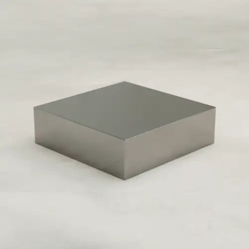 The Forge Steel Large Square Plinth, a metallic rectangular block with industrial flair, sits on a light, textured surface and casts a subtle shadow to one side.