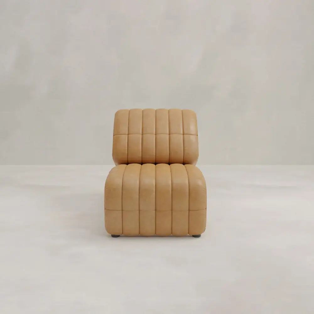 A modern Hillier Chair, armless and upholstered in premium vintage tan leather with vertical tufted lines, sits on a light floor against an off-white background.