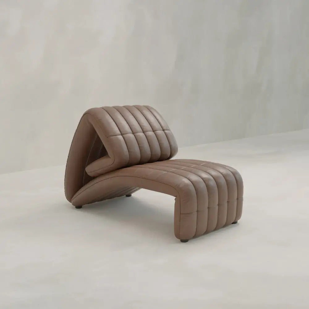 A Hillier Chair in premium vintage leather, with a curved wave design and vertical tufted lines, reclines for added comfort. It rests on a light floor beside a simple textured wall—an elegant touch for any modern lounge space.