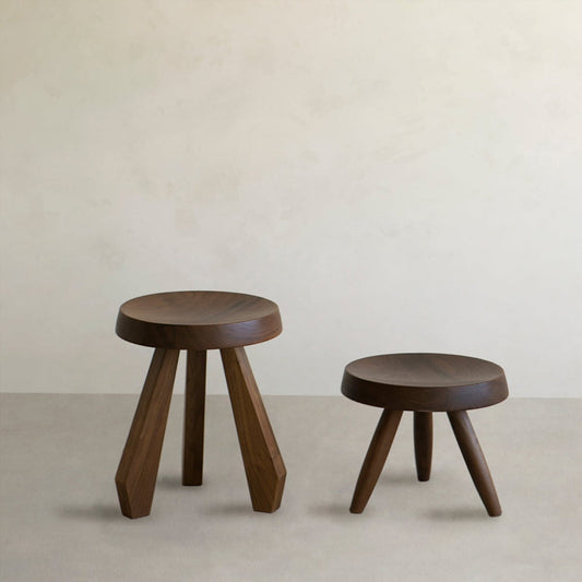 The Amara and Athena Stool Set Rental features two hand-carved wooden stools with round seats and tripod bases; one is taller than the other, both positioned on a neutral floor against a light beige wall.
