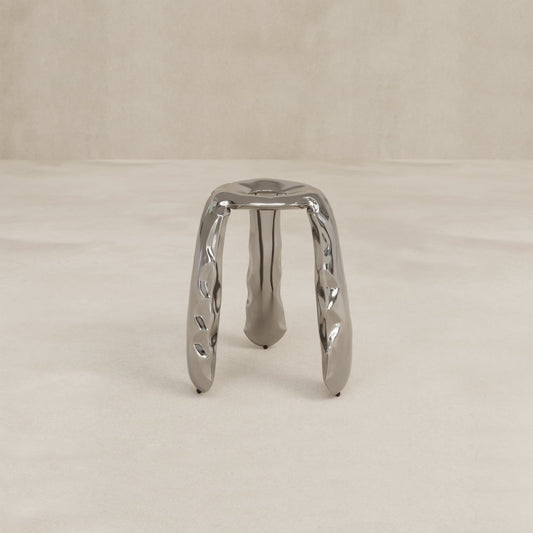 The Araknid Stool Large Rental, featuring three thick sculptural legs and sturdy metallic construction, stands on a light, textured surface against a plain beige background.