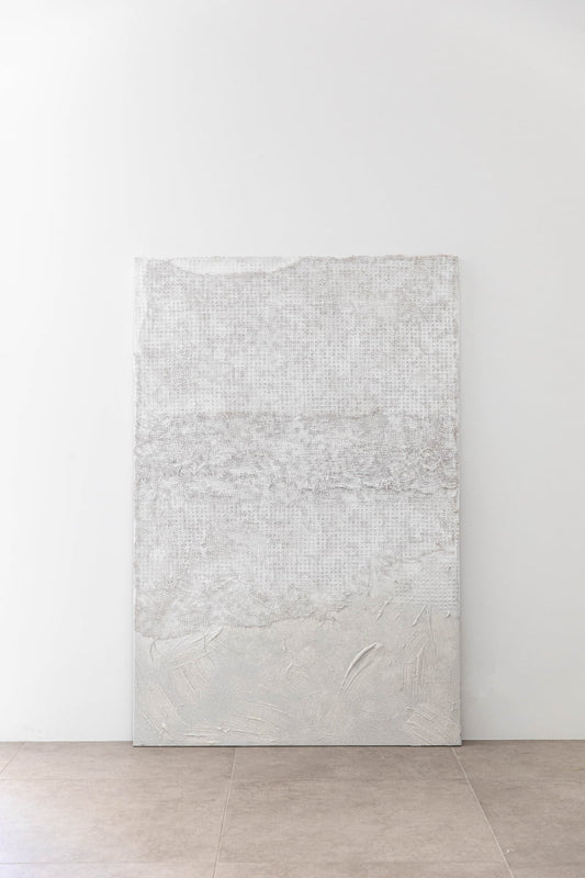 Art By Suzette Newman, a textured handmade abstract white canvas, rests against a white wall on a tiled floor. The artwork displays subtle patterns and raised areas for a layered, tactile appearance.