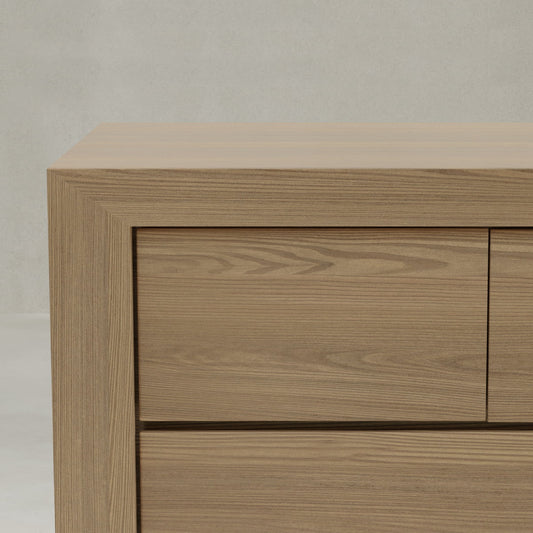 A close-up of the corner of the Ashford Large Dresser, a modern 12-drawer piece made from oak with clean lines, smooth surfaces, and visible wood grain, set against a plain light gray wall.