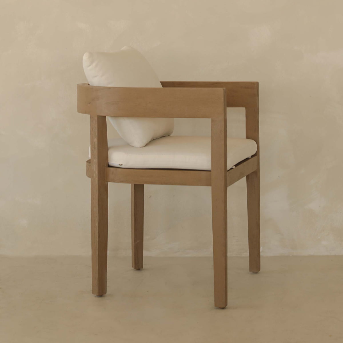 The Astier Outdoor Dining Chair features a minimalist wooden design with a square frame and armrests, plus white seat and back cushions, showcasing durable artisanal artistry against a light beige wall.