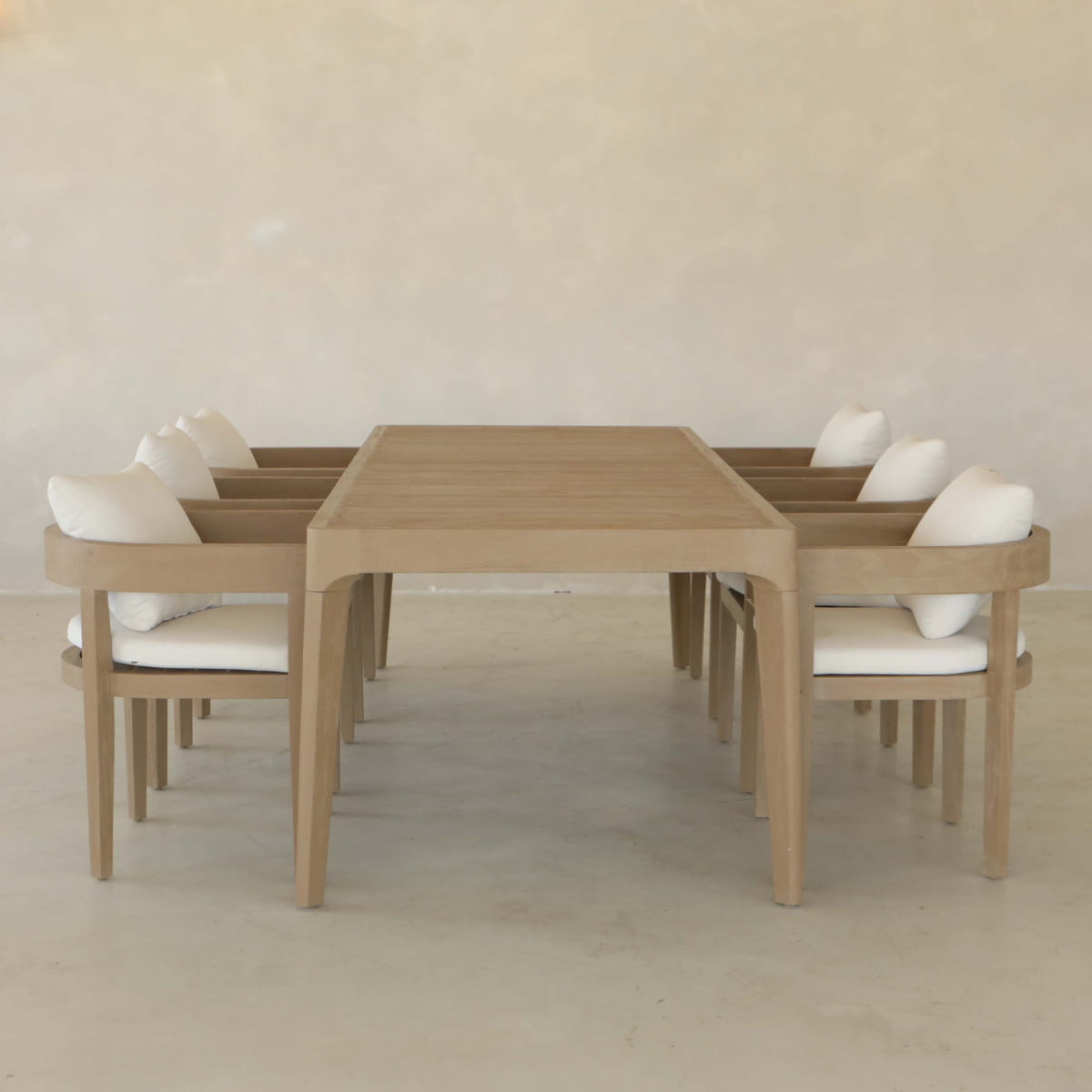 The Astier Outdoor Dining Table Set features a modern wooden table inspired by French Modernist design, with eight matching chairs—each with a white cushion and pillow—arranged in a minimalist room with light floors and walls.