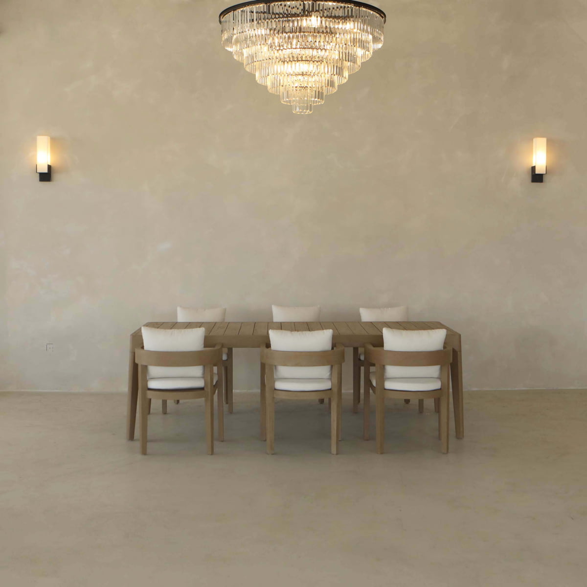 A minimalist dining room showcases the Astier Outdoor Dining Table Set, featuring a wood-inspired table with six white-cushioned chairs, two wall sconces, and a crystal chandelier against a light beige wall.