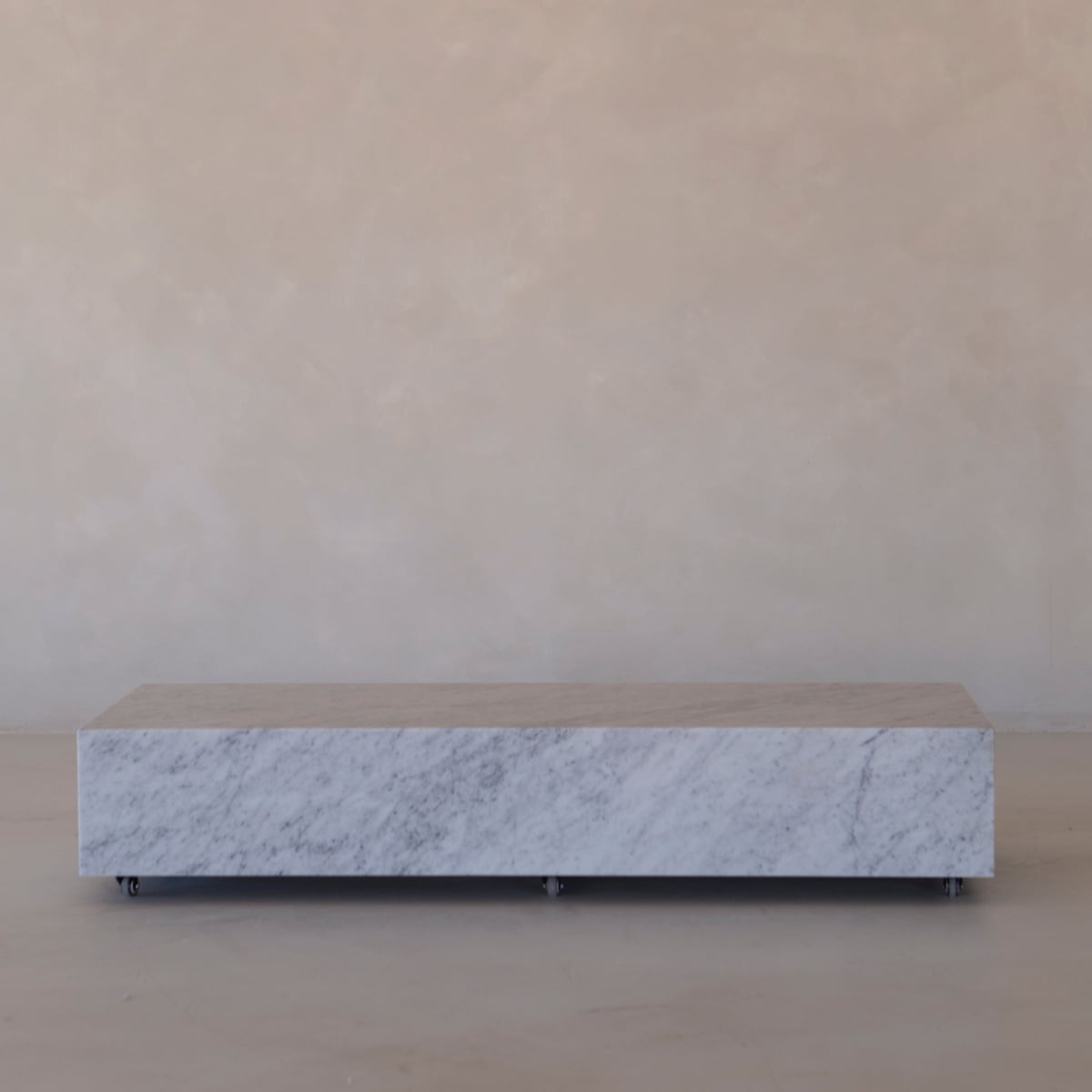 The Aurora Marble Plinth, a rectangular table made from Carrara marble with subtle gray veining and smooth-rolling wheels, is placed against a beige textured wall and floor.