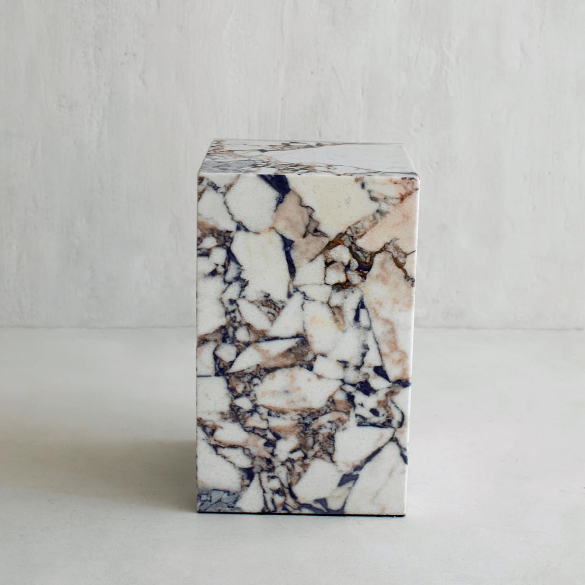 The Turkish Calacatta Plinth Tall is a rectangular white marble block with brown, beige, and dark gray veining, resting on a light surface against a textured light gray wall.