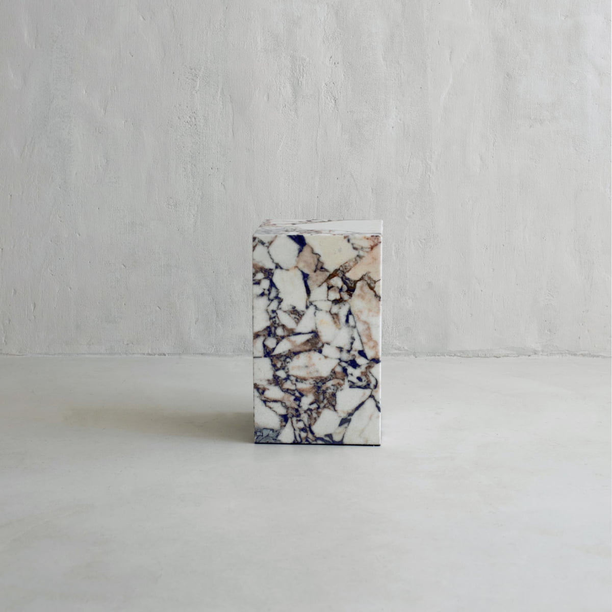 The Turkish Calacatta Plinth Tall, featuring a white base with brown, black, and gray veining, stands on a smooth light gray floor against a textured pale wall.