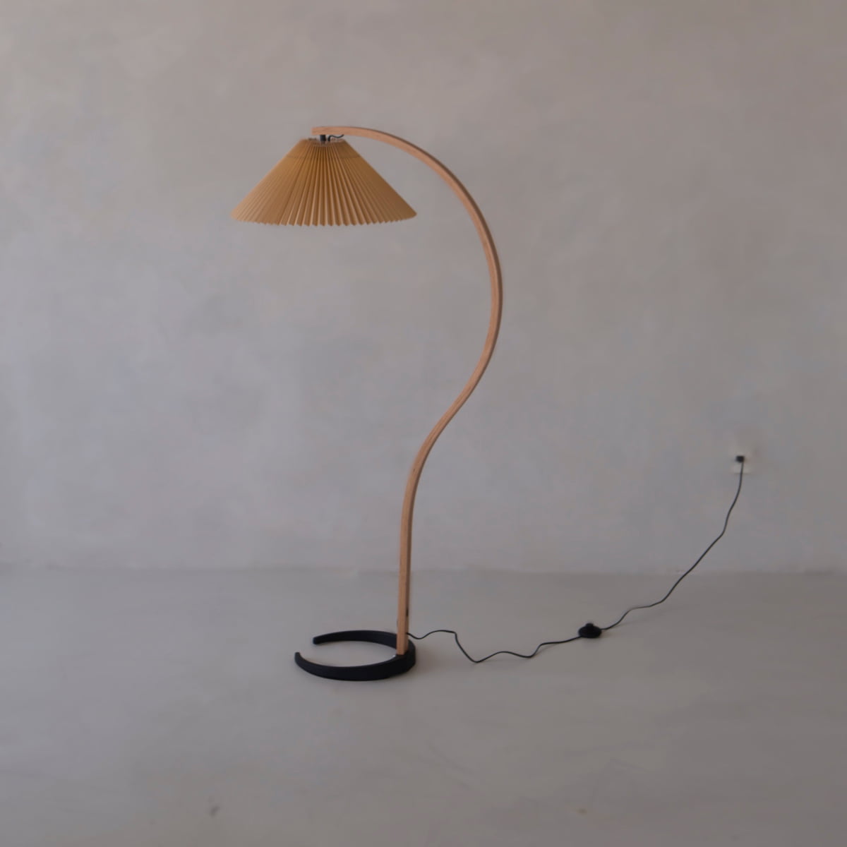 The Bailey Lamp features a curved wooden stand, pleated beige lampshade, and black circular base for modern elegance against a light gray wall, with its power cord discreetly plugged in.