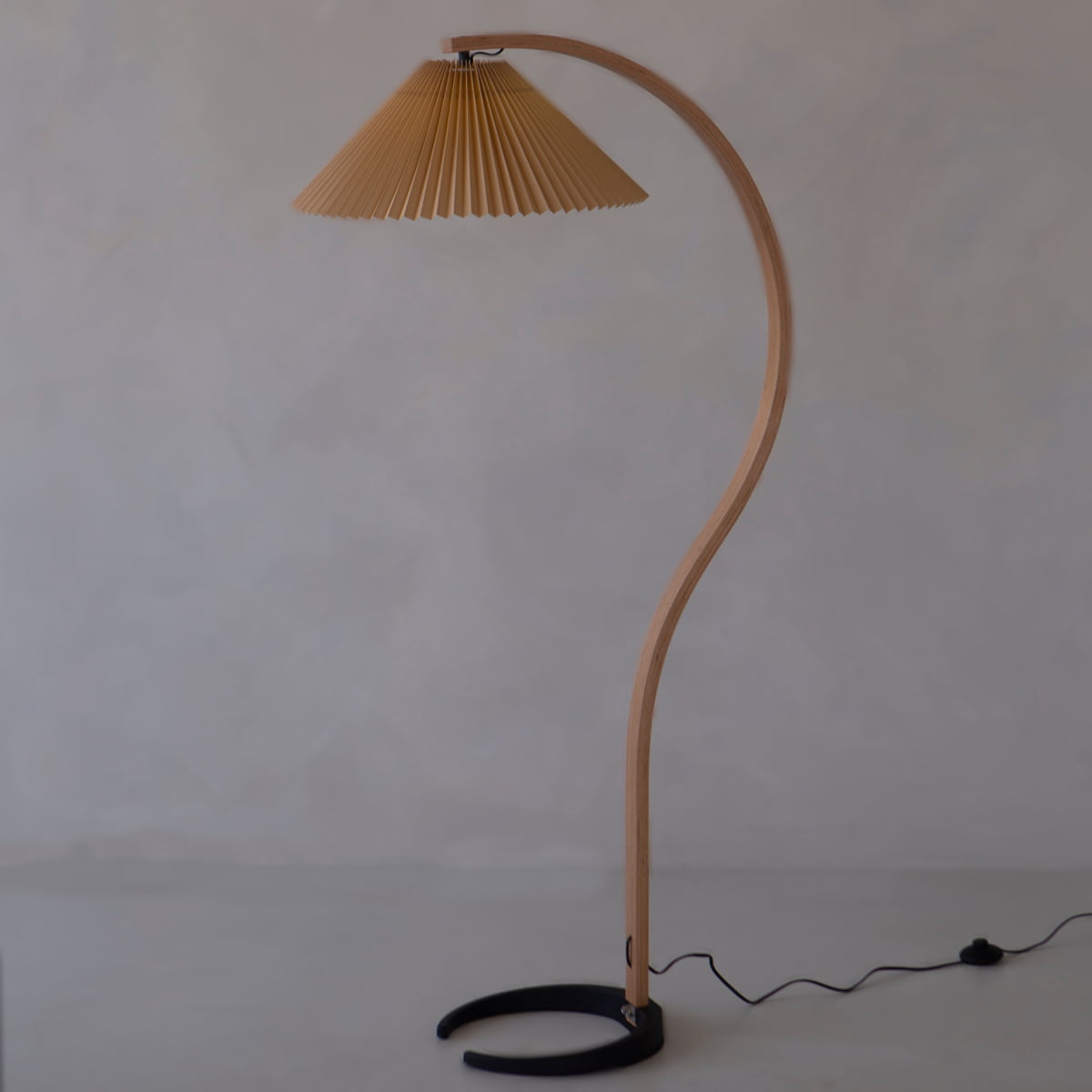 The Bailey Lamp features a curved wooden stand, pleated brown shade, and black semicircular base, bringing modern style to any light-colored floor and neutral wall. A black cord extends from the base for a sleek finishing touch.