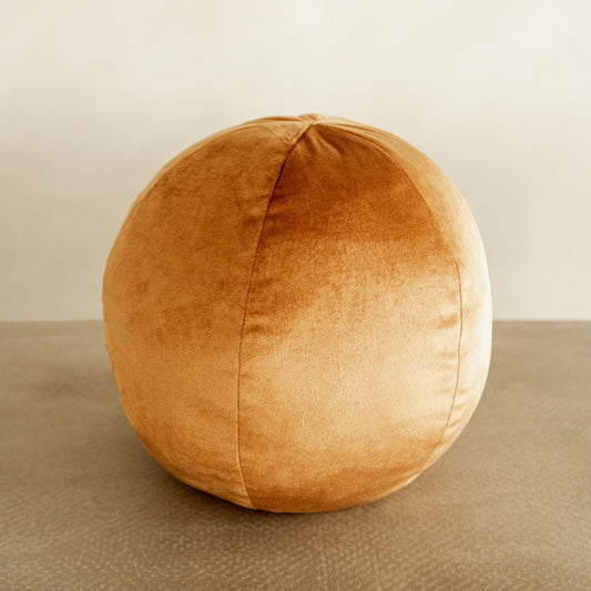 The Ball Pillow, a round tan bouclé velvet accessory, rests on a textured gray surface with a neutral beige background, adding a stylish decorative accent.