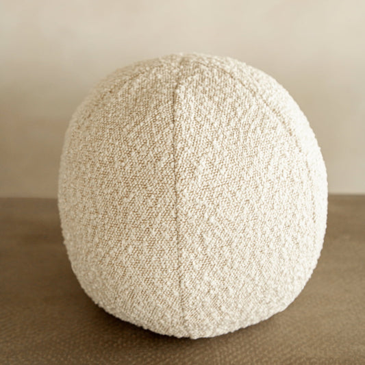 The Ball Pillow, featuring a textured off-white oval design with a vertical seam, rests on a smooth brown surface against a neutral background, serving as an elegant decorative accent.