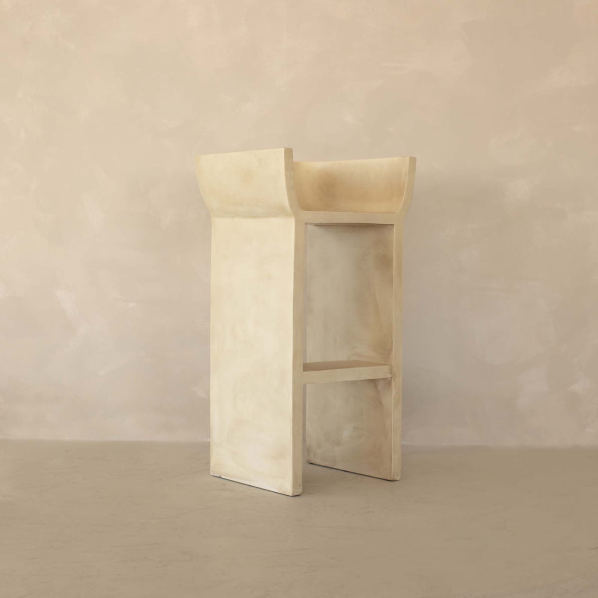 The Barbican Stool, featuring a modern minimalist Brutalist design with clean lines and an open rectangular form, stands on a neutral floor against a textured wall, exuding urban elegance.