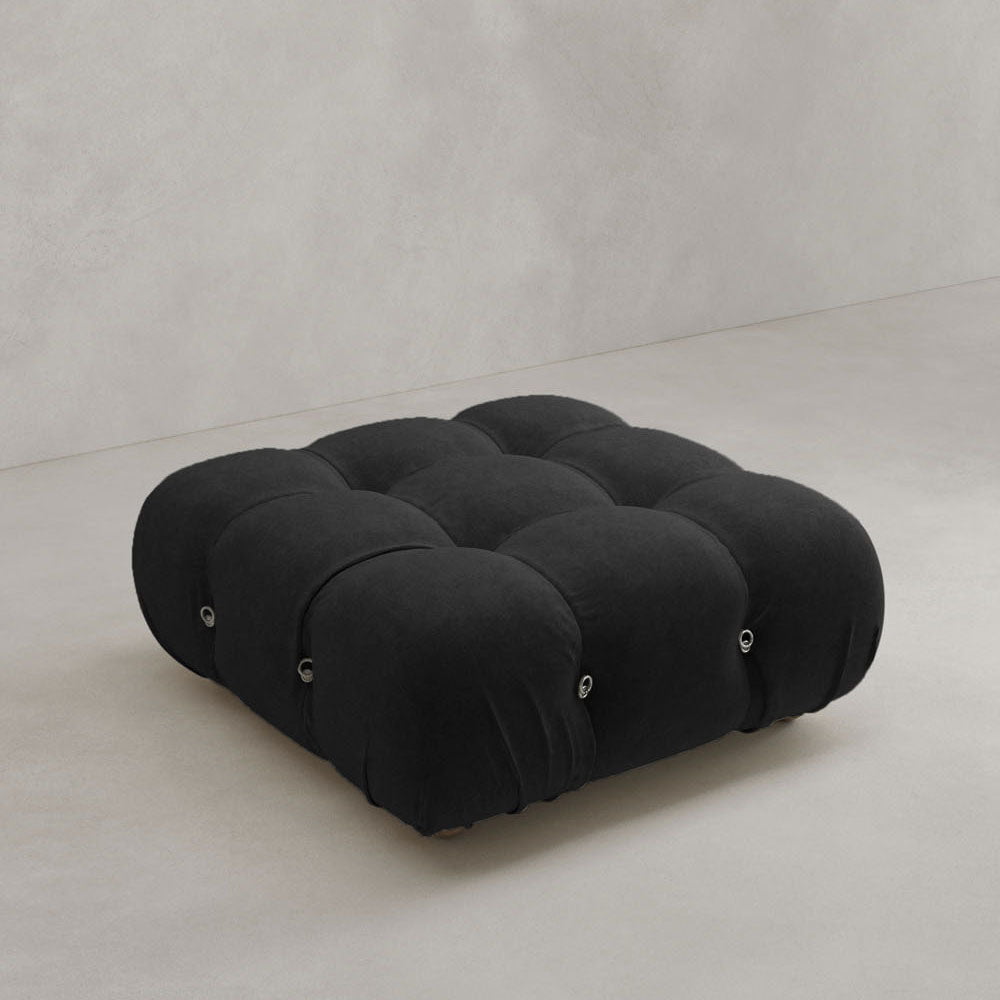 The Belmont Ottoman, a large, square black piece with tufting and rounded edges, features plush fabric and a modular design for a modern feel. It sits on a light gray floor against a plain, light wall, evoking an iconic classic style.