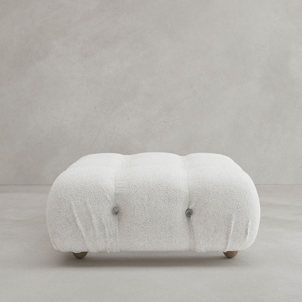 The Belmont Ottoman is a low, square piece with tufted, soft white textured fabric, rounded edges, and a modular design, shown against a plain light gray background.