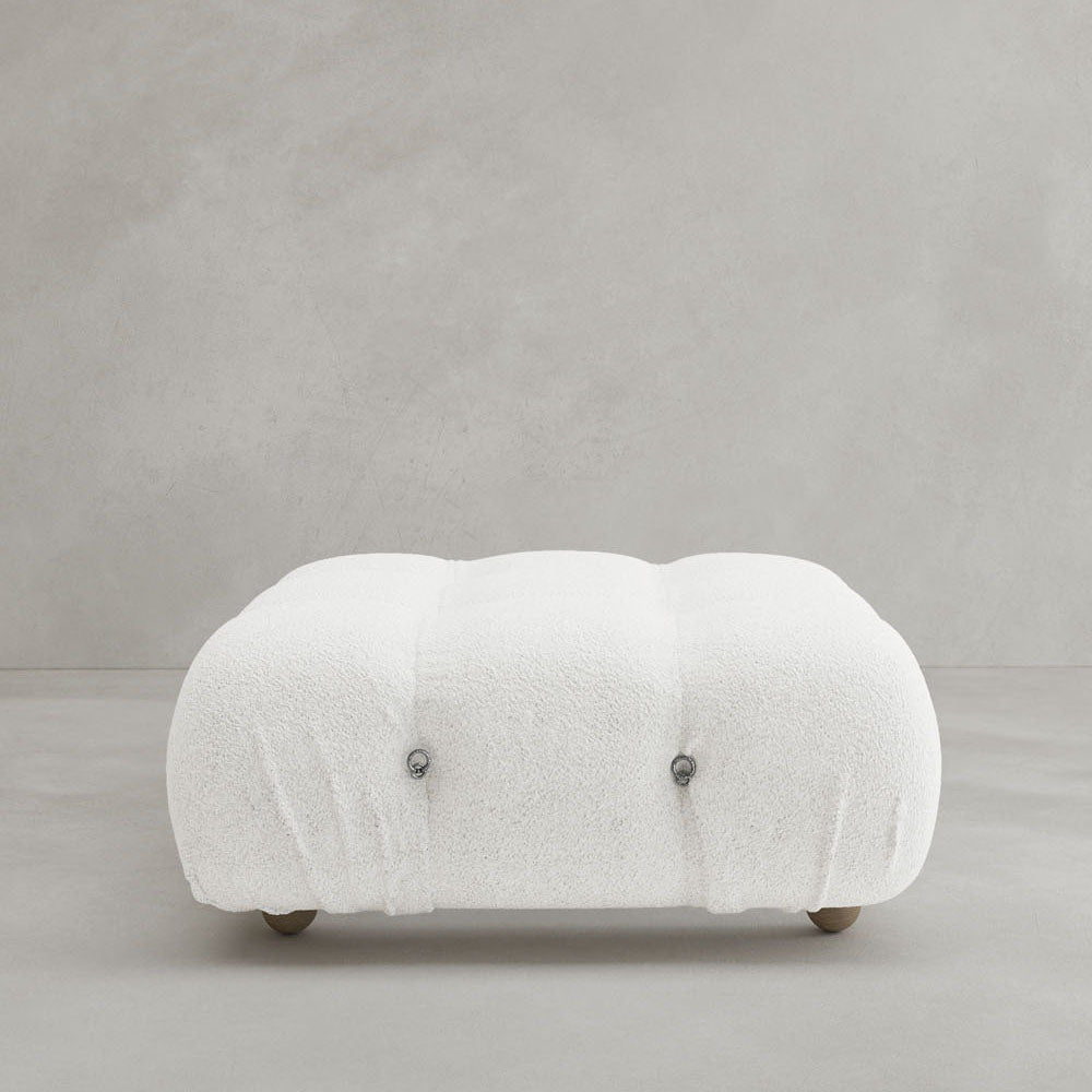 The Belmont Ottoman features a modern rectangular design in white with a plush, tufted top and short wooden legs, reflecting iconic classic style against a plain light gray background.