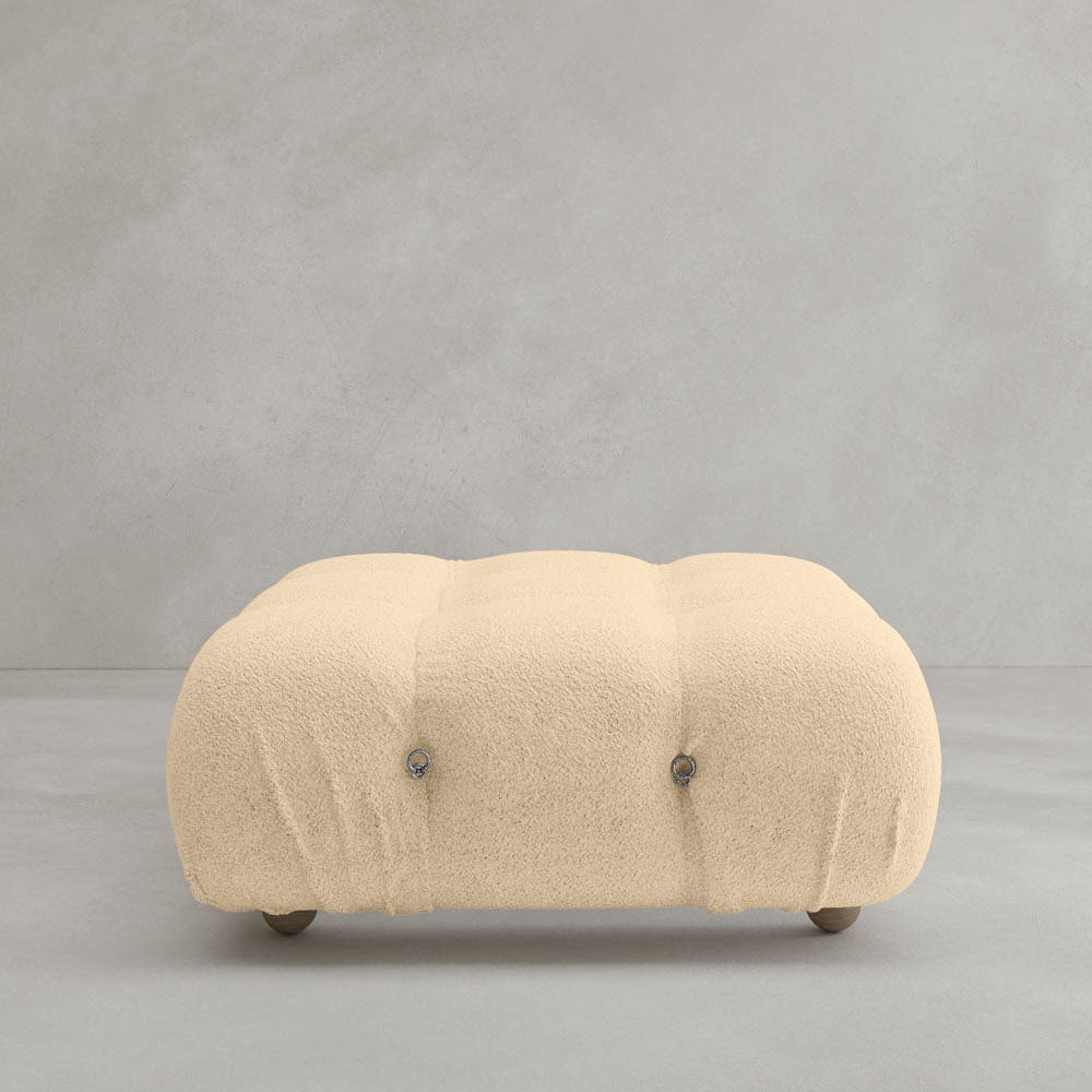 The Belmont Ottoman features a beige, tufted design with soft, textured fabric and modular style. It has rounded edges, short hidden legs, and sits on a light gray floor against a matching gray wall.