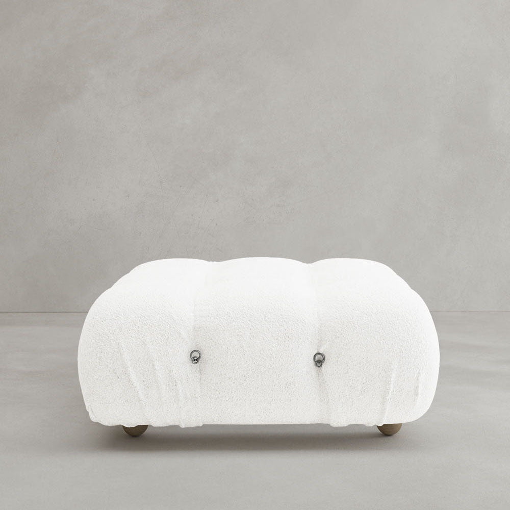 The Belmont Ottoman is a low, rectangular piece with tufted white, textured upholstery and small round feet, inspired by classic modular design, shown against a plain light gray background.