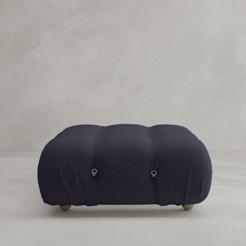 The Belmont Ottoman, featuring a dark gray, rounded form and soft textured surface, rests on small wooden legs with two metal ring accents. Its modular design adds charm to any light-colored floor and plain light gray wall setting.