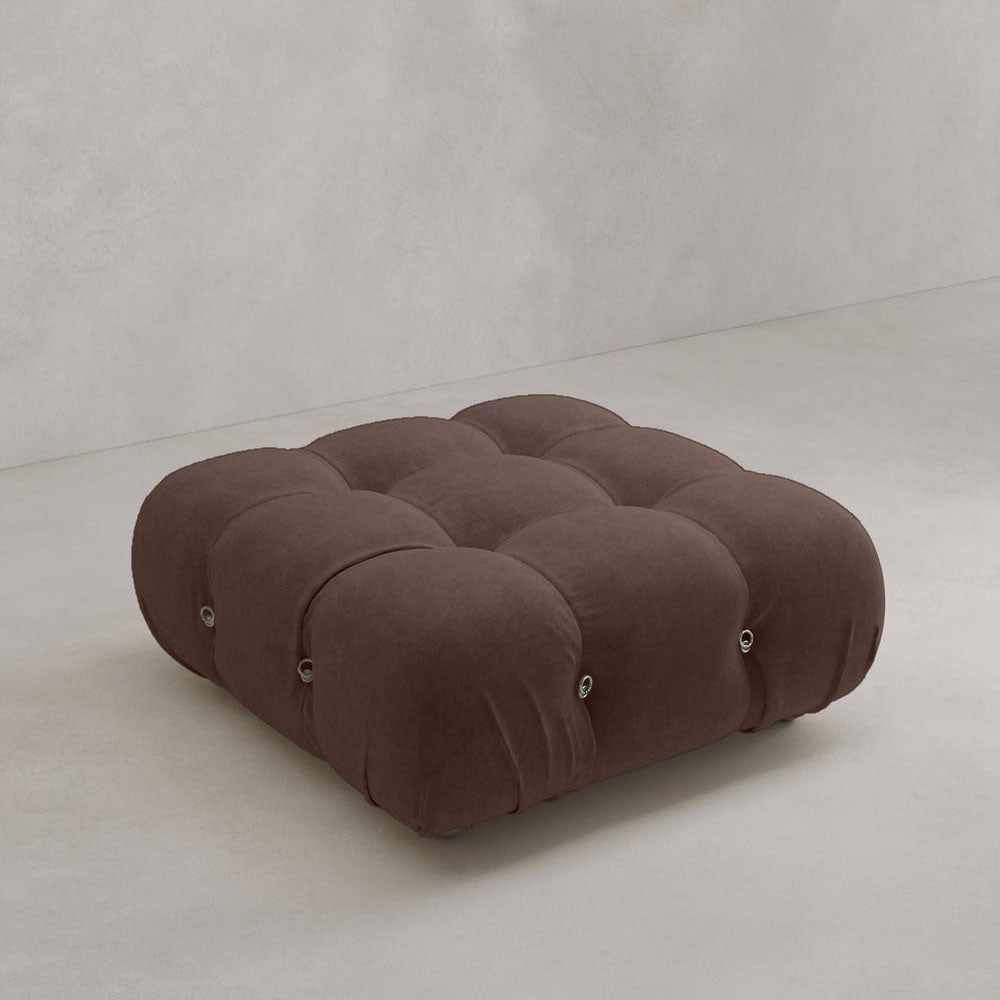 The Belmont Ottoman, a low square piece in dark brown suede with tufted buttons and rounded edges, brings classic style to any minimalist space.