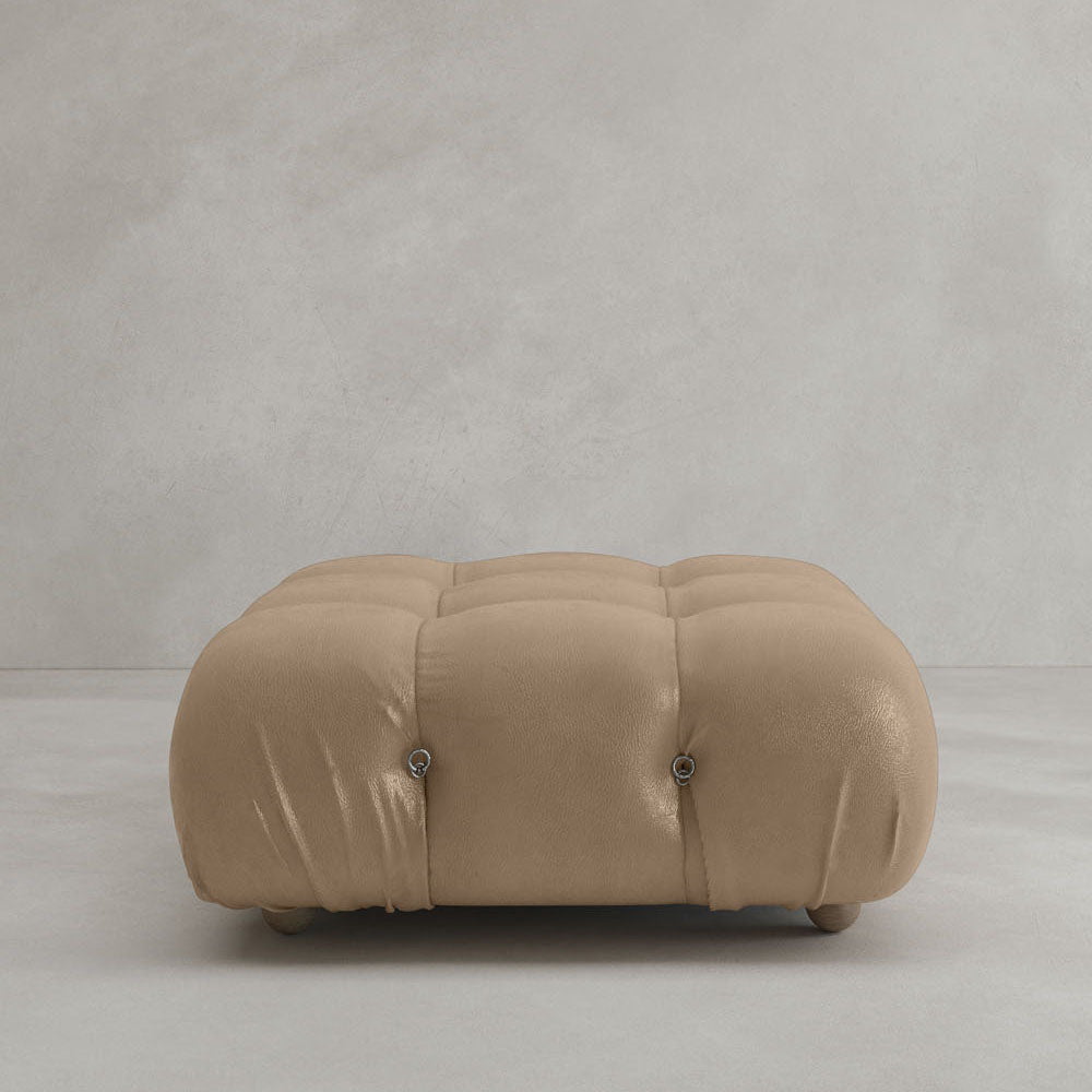 The Belmont Ottoman, featuring a tan tufted design and soft cushioning, is placed on a light gray floor against a plain, light-colored wall.