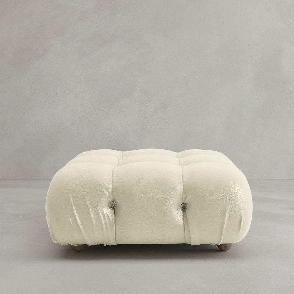 The Belmont Ottoman is a low, square, cream-colored piece with button tufting and modular design, shown on a smooth light gray floor against a plain gray wall.