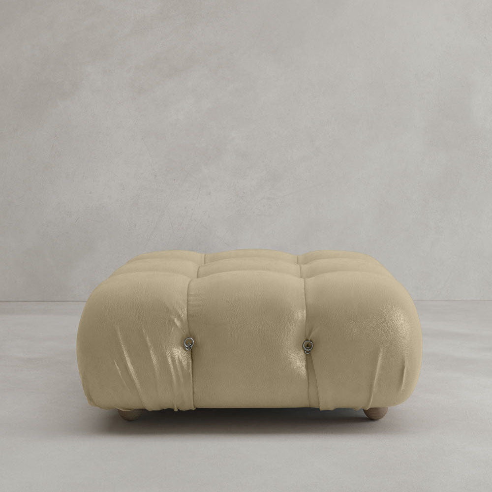 The Belmont Ottoman, in beige with tufted detailing and a soft cushioned top, sits on a light gray floor against a plain, light-colored wall.