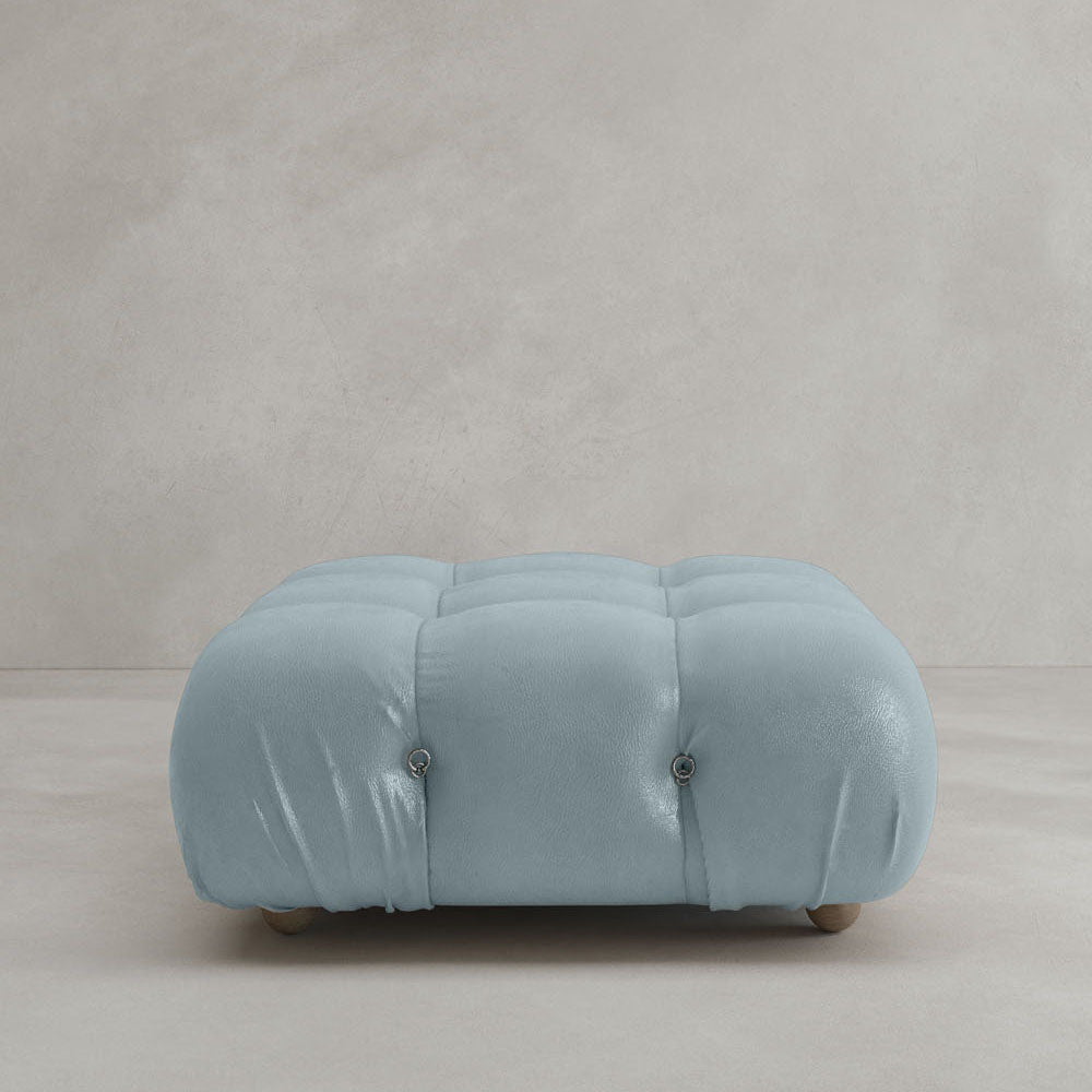 The Belmont Ottoman is a large, square, light blue piece with a tufted cushioned top and four small wooden feet, featuring modular design and displayed on a plain light gray background.