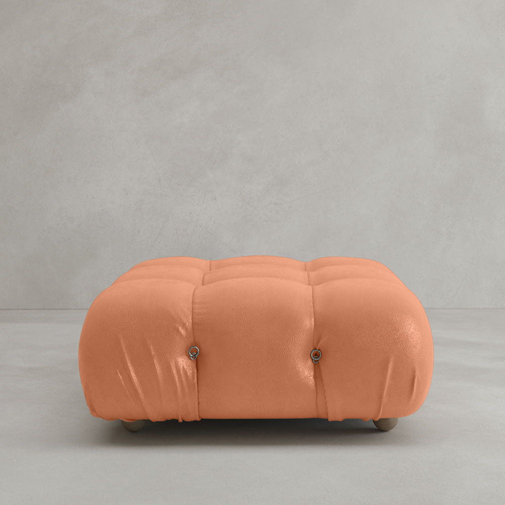 The Belmont Ottoman, a square tufted piece in peach with rounded edges and small wooden legs, sits on a light gray floor against a plain gray wall, adding an iconic, classic accent ideal for modular interiors.