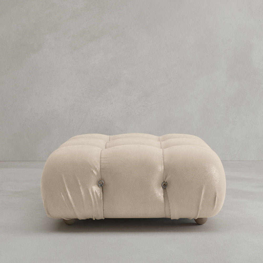 The Belmont Ottoman is a square, tufted, cream-colored piece with soft, rounded edges, resting on a light gray floor against a plain gray wall—its iconic look pairs perfectly with cloud-like sofas or modular designs.
