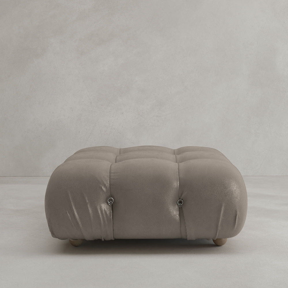The Belmont Ottoman features a square, tufted design with soft light brown leather upholstery and four short round wooden legs, showcasing classic style on a light gray floor against a textured light gray wall.