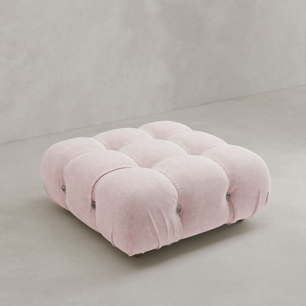 The Belmont Ottoman, a large square blush pink piece with rounded edges and a modular tufted design, sits on a light grey floor against a plain grey wall.