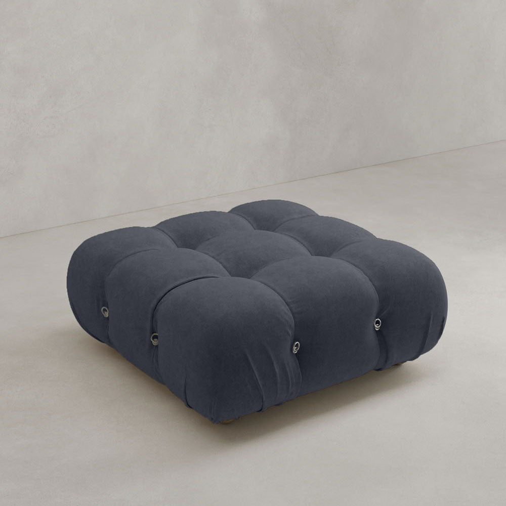 The Belmont Ottoman is a modern, dark grey, tufted piece with a plush cushioned surface and modular design, shown on a light floor against a neutral wall.