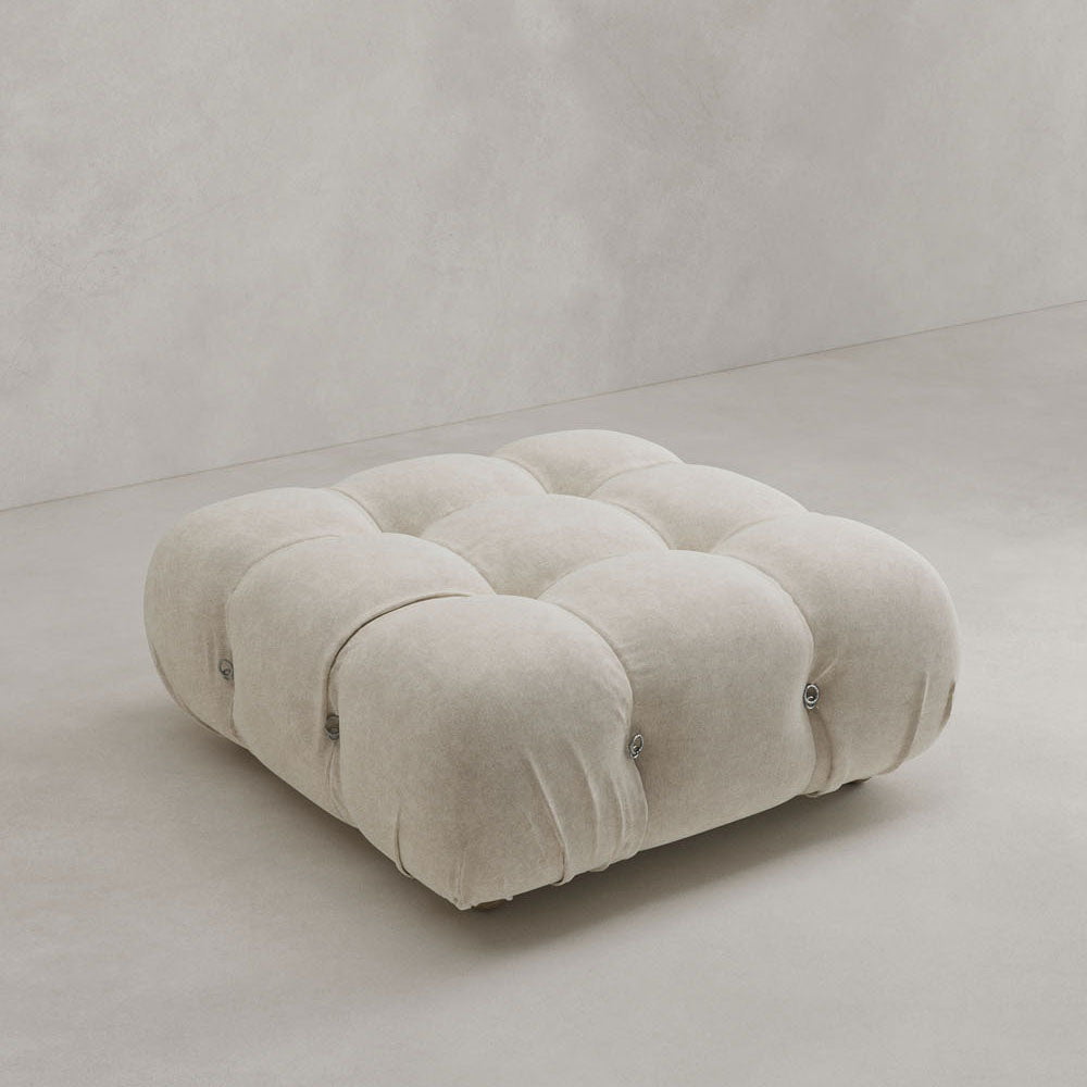 The Belmont Ottoman, a square tufted ottoman in light beige with a plush texture, sits on a smooth light floor against a neutral wall—an iconic classic that complements any modern space.