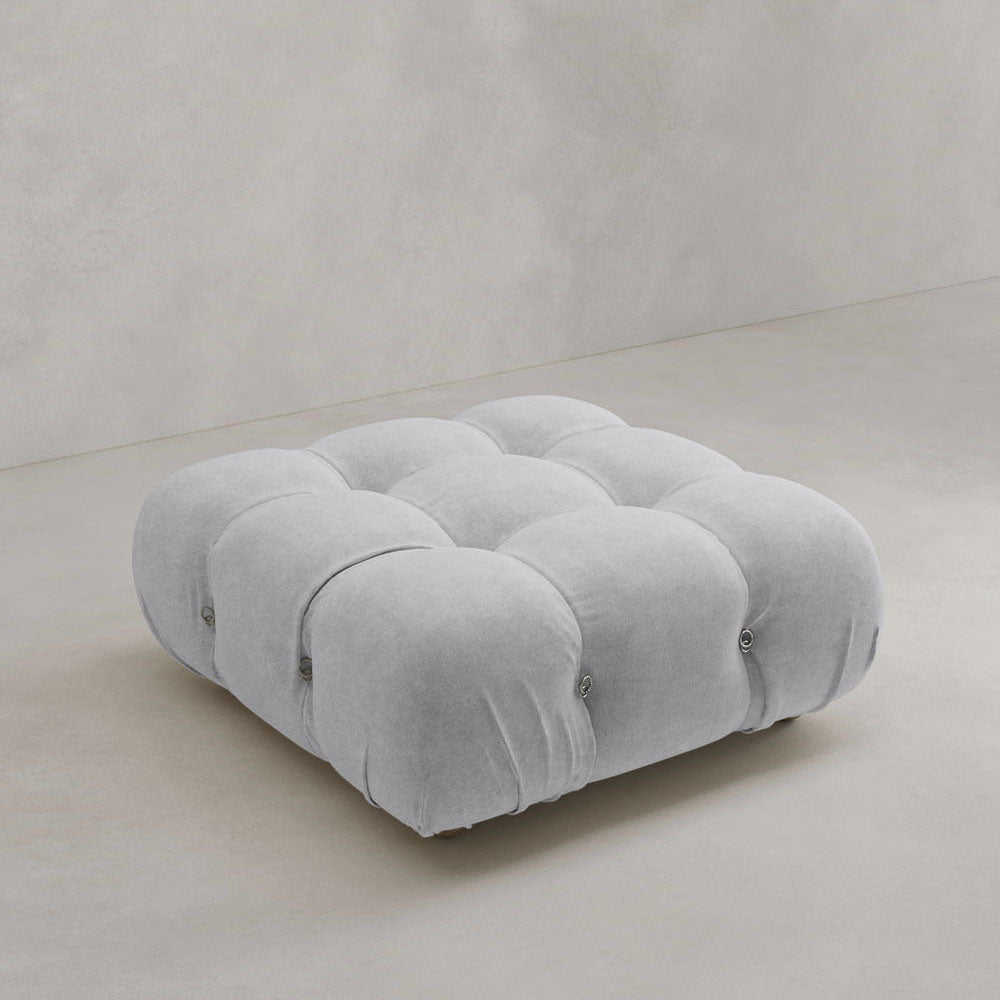 The Belmont Ottoman, featuring a square tufted design and soft, plush light gray upholstery, sits on a smooth, light-colored floor against a plain wall.