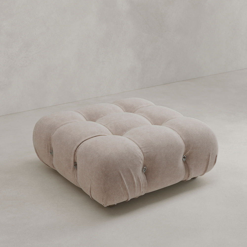The Belmont Ottoman features a square, tufted design in soft light beige upholstery, sitting on a neutral floor against a matching wall—ideal for complementing modular setups or iconic classic furniture.