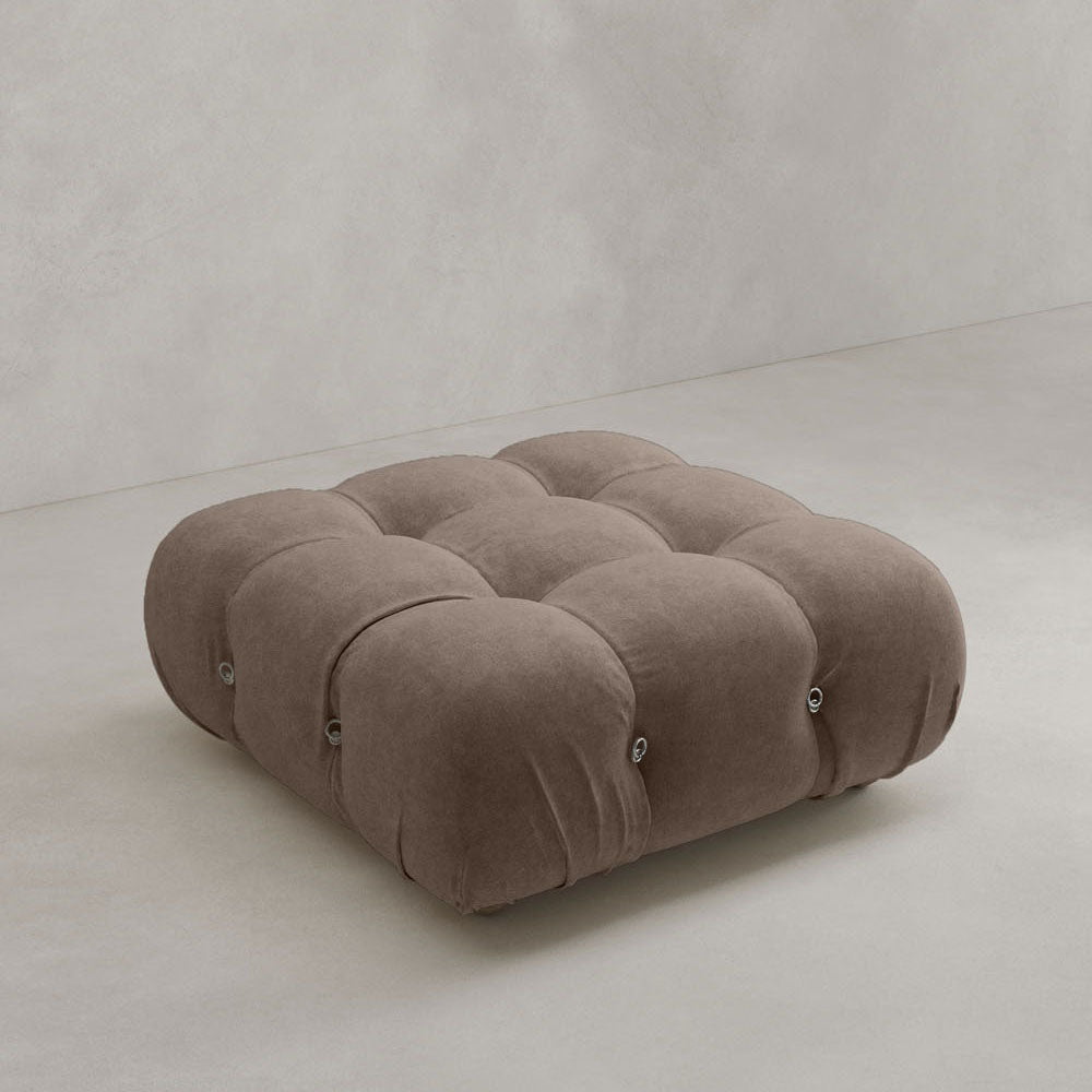 The Belmont Ottoman is a large, square, tufted piece with a modular design, upholstered in soft brown fabric. It features rounded edges and a plush, cushion-like look, set on a light gray floor against a plain light-colored wall.