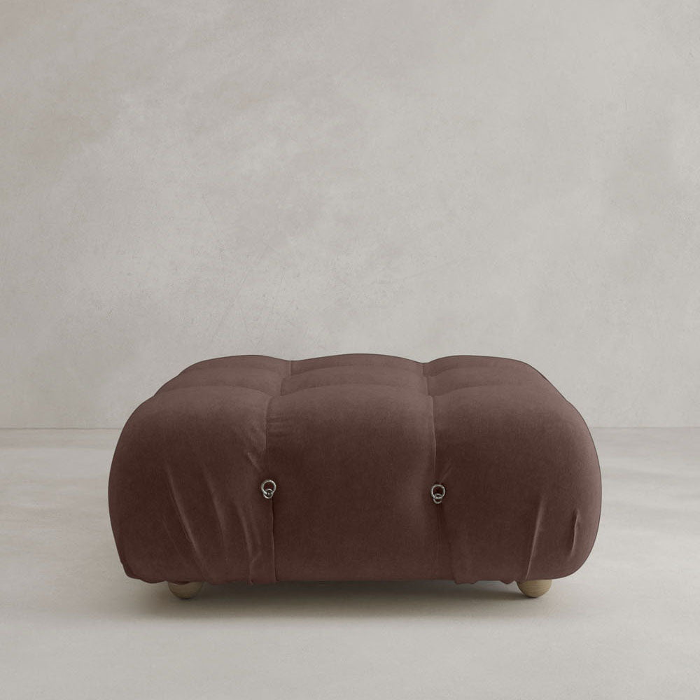 The Belmont Ottoman—a large, square, brown piece with tufted details, rounded edges, and short wooden feet—sits on a light floor against a grey wall, making it ideal for classic or modular design setups.