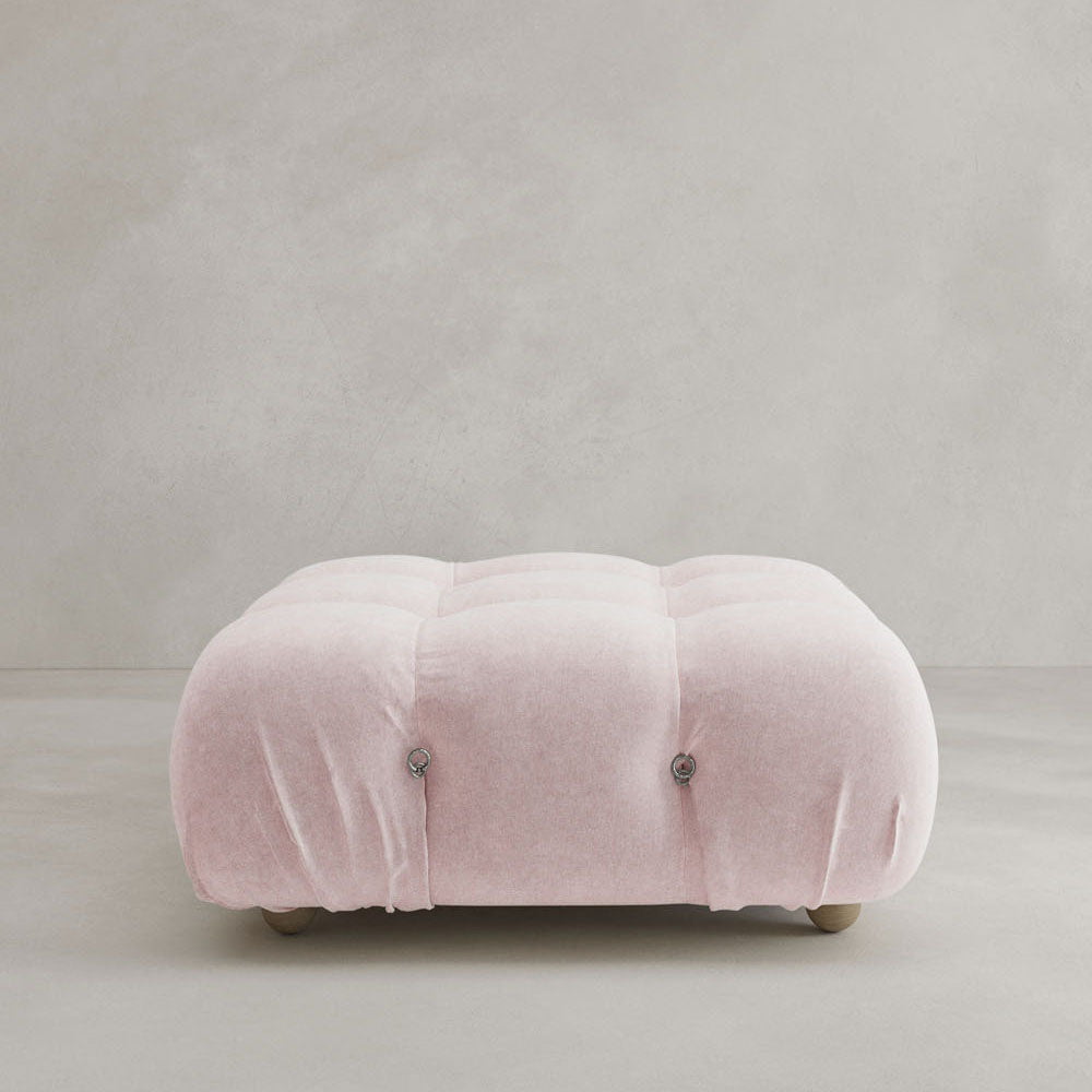 The Belmont Ottoman is a light pink, square, tufted piece with rounded edges and button details on small wooden legs, shown against a plain light gray background—a modular design that pairs perfectly with any classic or cloud-like sofa.