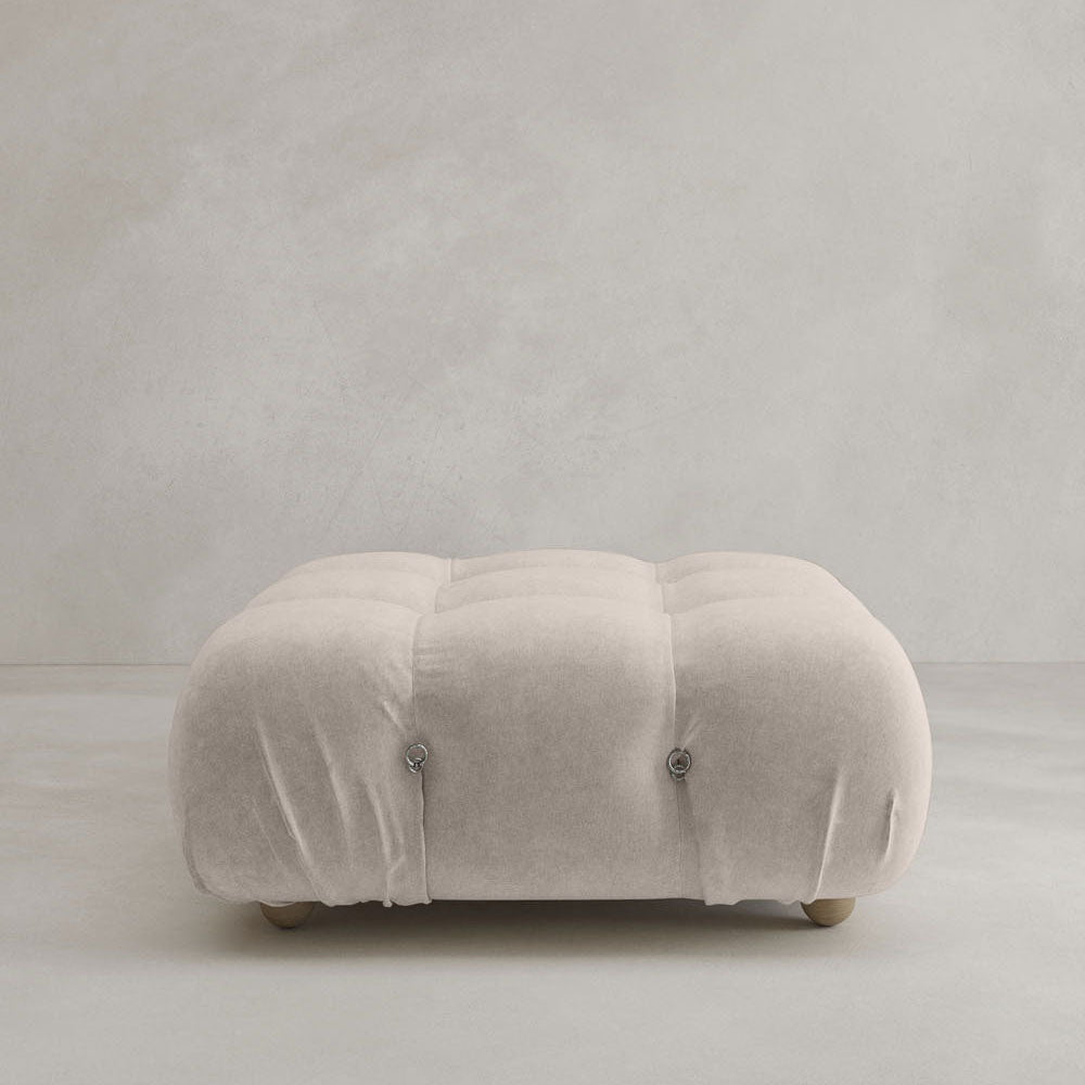 The Belmont Ottoman, a square, tufted piece upholstered in light beige fabric with refined button detailing and classic style, is set against a plain light gray background.