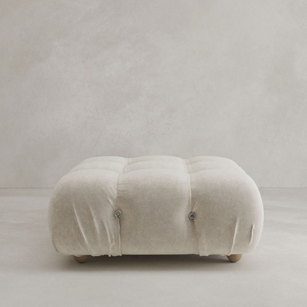 The Belmont Ottoman is a plush, square, light beige seat with tufted buttons and short wooden legs, offering a classic look that complements modular furniture against light-colored walls and floors.