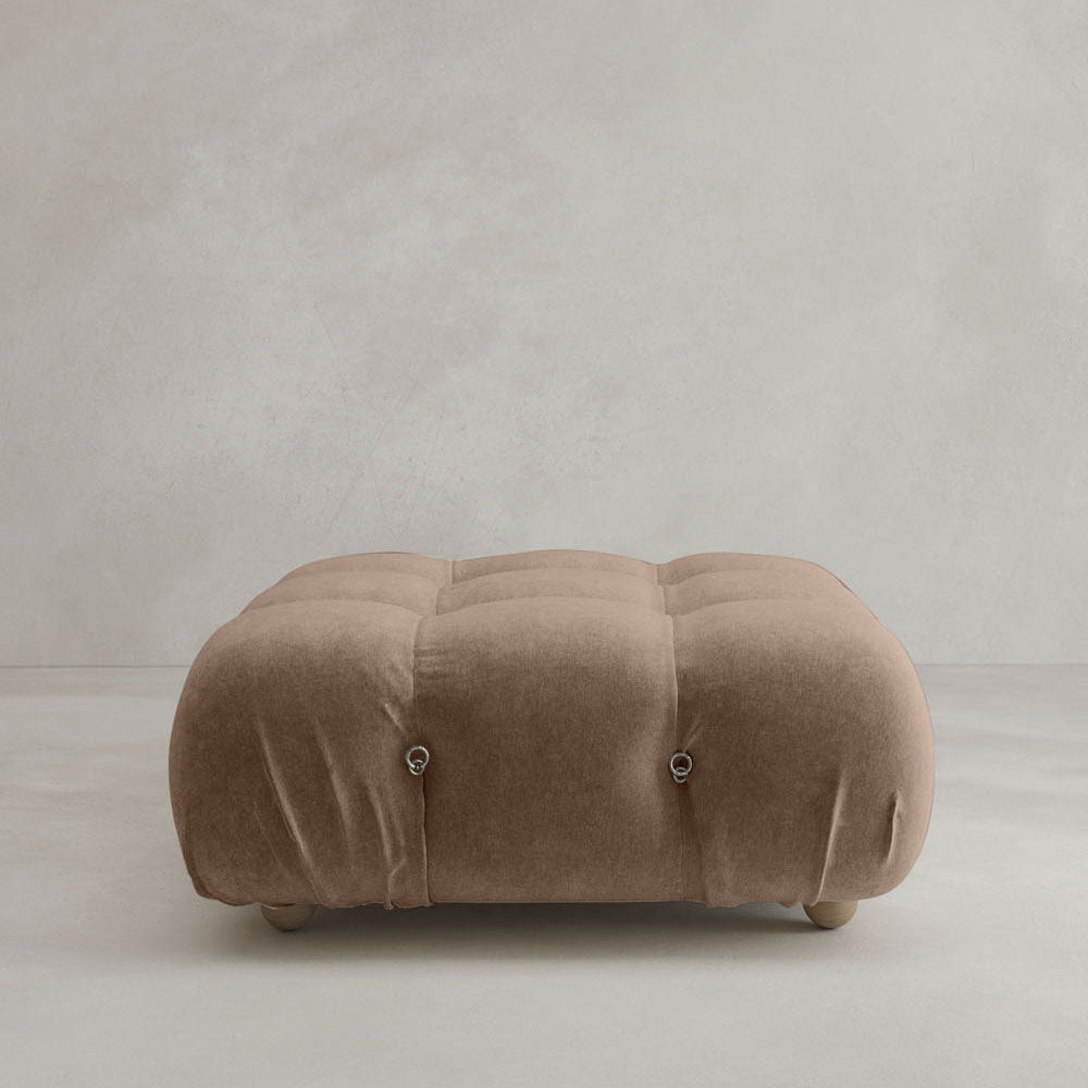 The Belmont Ottoman, a soft square piece with light brown suede-like fabric and modular-inspired tufting, rests on a light floor against a neutral textured wall.